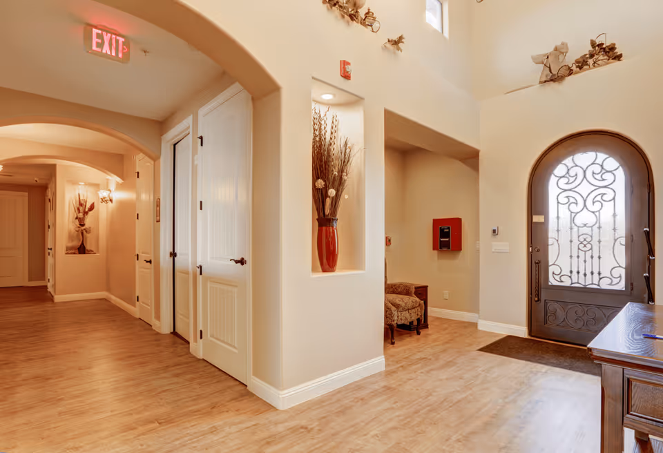 Interior view of a senior living facility hallway with wooden flooring, beige walls, white doors, and decorative niches containing vases with dried plants. There is an ornate glass front door on the right side and an exit sign hanging from the ceiling on the left side.