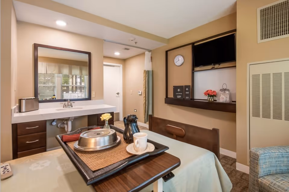 A senior living bedroom featuring an overbed meal tray, sink and mirror, wall-mounted TV, and seating.