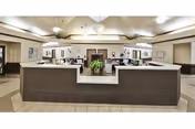 A spacious nursing station area with a large, dark brown reception desk in the center. The desk has a white countertop and a green potted plant placed in the middle. The background shows office equipment, chairs, and doors leading to other rooms. The ceiling has recessed lighting and decorative light fixtures.