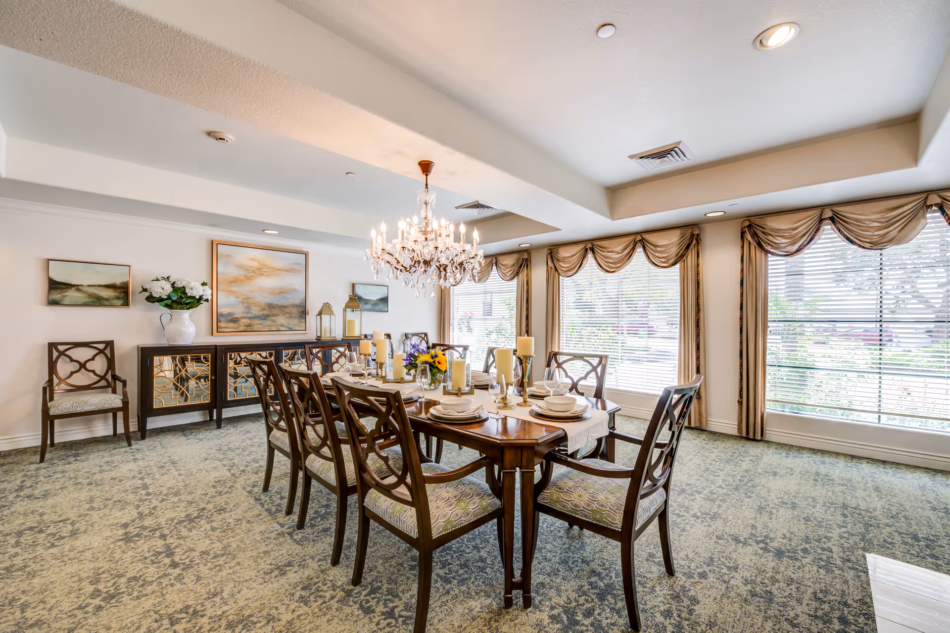 A well-lit dining room with a long wooden table set for eight people. The table is decorated with candles and a floral centerpiece. There are eight wooden chairs with patterned cushions around the table. A large chandelier hangs above the table. The room has large windows with beige curtains and blinds, and a sideboard with decorative items and paintings on the wall behind it.