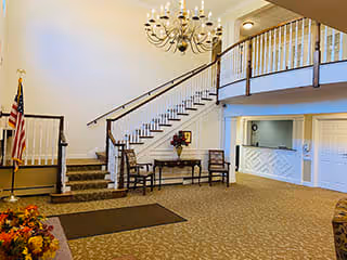 Spacious senior living facility lobby with a staircase leading to an upper floor, a small table with chairs and a flower arrangement, an American flag, and a reception area with a counter.
