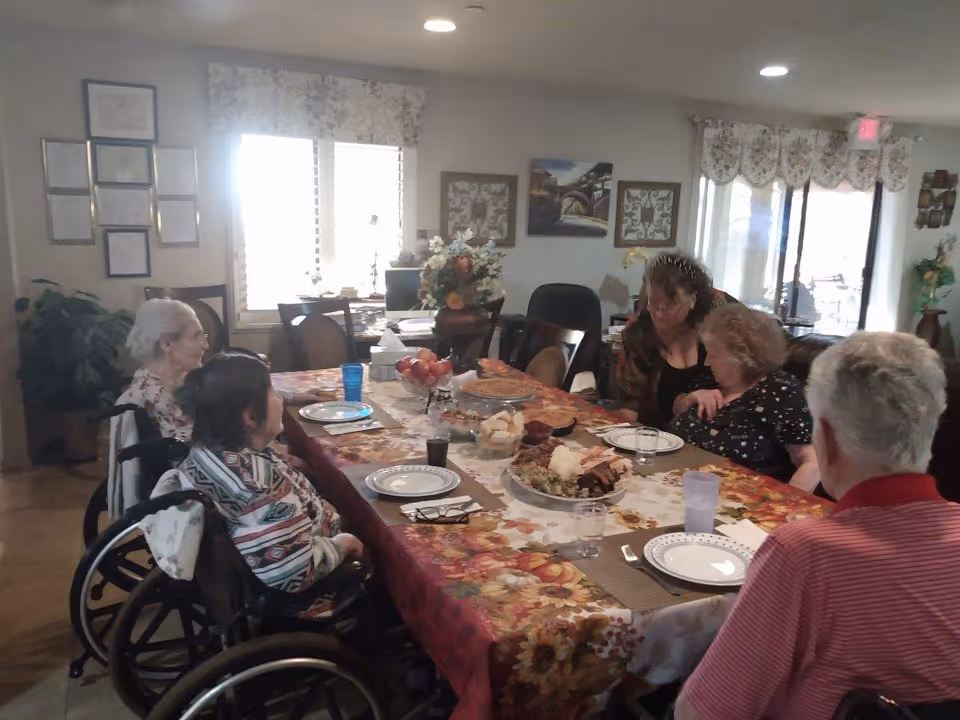 A group of elderly people sitting around a dining table covered with a floral tablecloth, with plates and glasses set for a meal. The room is well-lit with windows and decorated with framed pictures and a flower arrangement on the table.