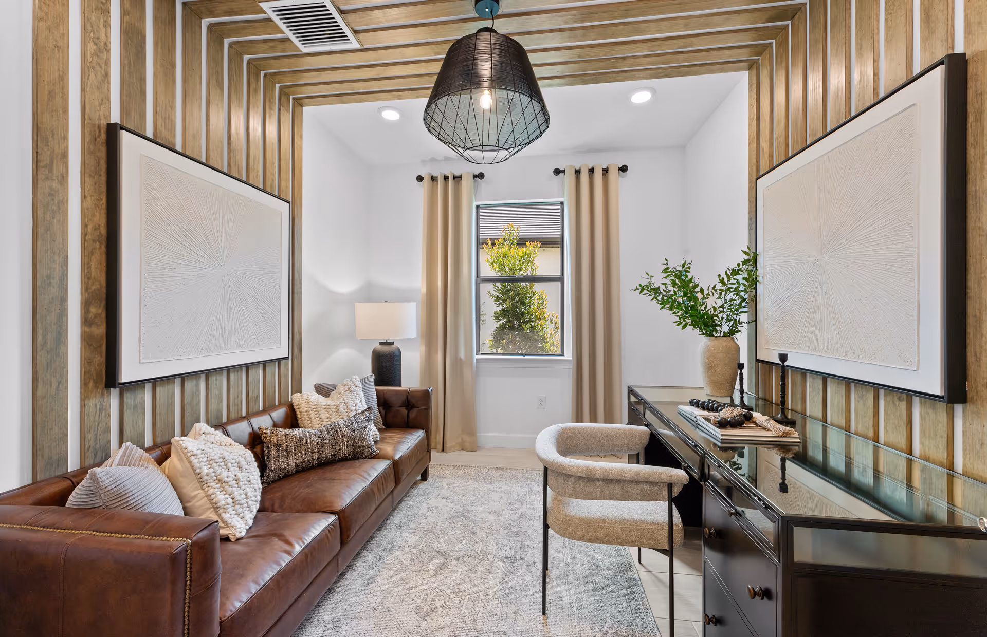 A modern living room with a brown leather sofa adorned with multiple textured pillows, a black desk with a glass top, a beige cushioned chair, and a large window with beige curtains. The walls feature vertical wooden paneling with two large abstract artworks, and a black pendant light hangs from the ceiling.