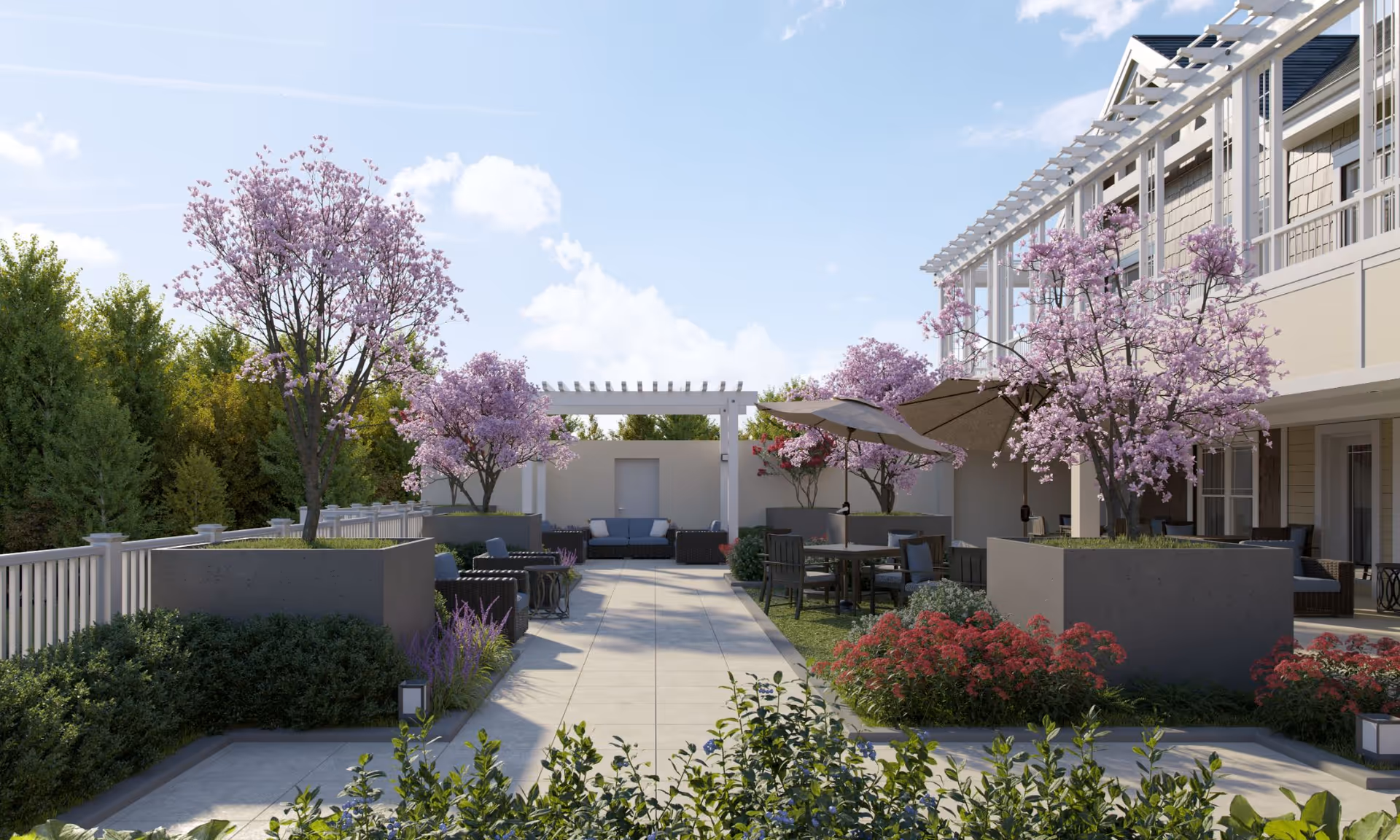Outdoor patio area at Sunrise of Livingston featuring a paved walkway, seating areas with chairs and tables, large planters with blooming pink trees, umbrellas for shade, and surrounding greenery under a blue sky with some clouds.