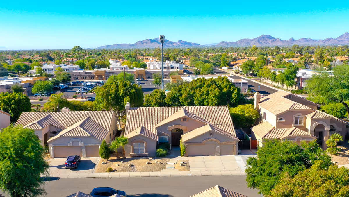 Aerial view of a suburban street lined with tile-roofed homes, trees, a nearby shopping area, and mountains in the distance.