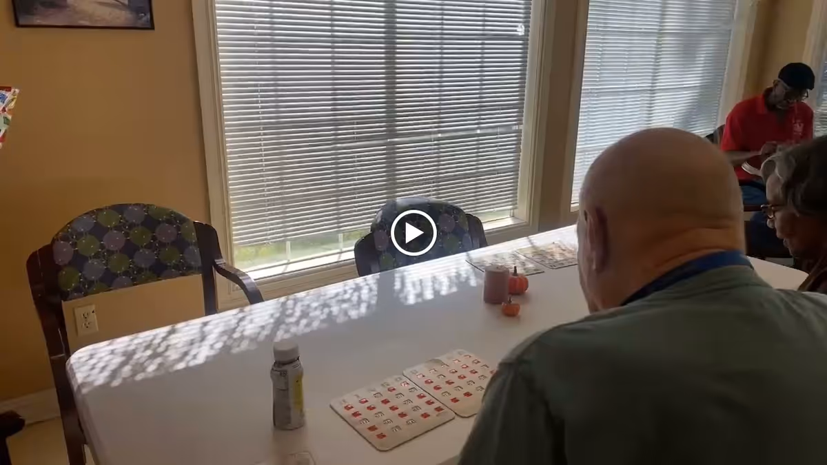 Two elderly individuals sitting at a white table playing a game with bingo cards. The room has large windows with blinds partially closed, allowing sunlight to cast shadows on the table. There are patterned chairs around the table and a small pumpkin decoration on the table.