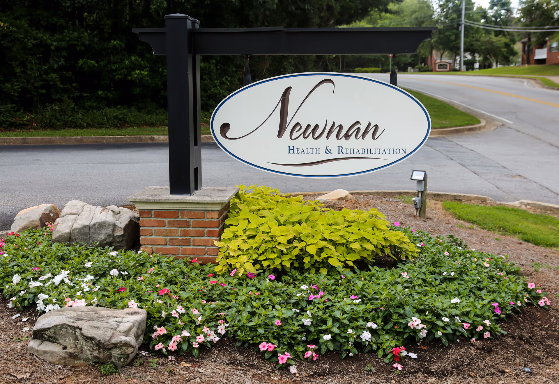 Entrance sign reading "Newnan Health & Rehabilitation" mounted on a post above a flower bed by the roadside.