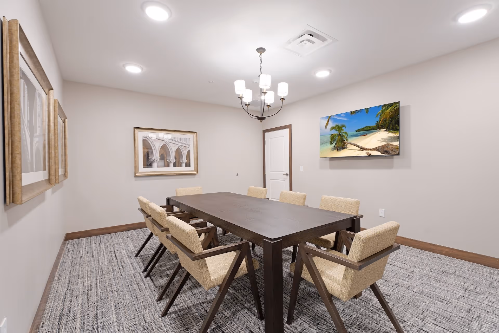 A modern meeting or dining room with a rectangular dark wood table surrounded by eight beige cushioned chairs. The room has light gray walls, a patterned carpet, a chandelier with six lights hanging from the ceiling, and recessed ceiling lights. On one wall, there is a flat-screen TV displaying a tropical beach scene, and on another wall, there are two framed pictures of architectural arches.