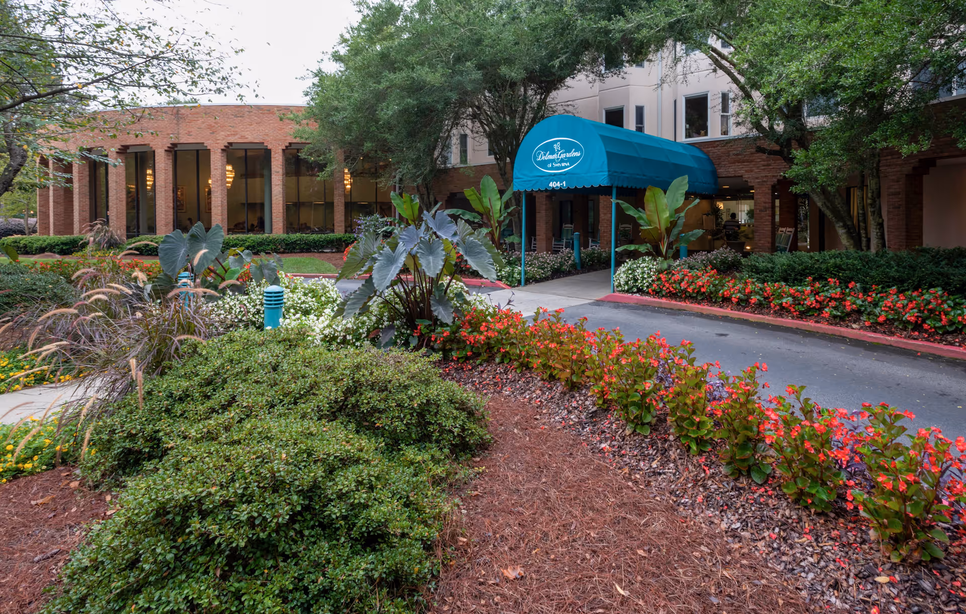 Entrance to Delmar Gardens of Smyrna facility with a blue canopy over the driveway, surrounded by well-maintained landscaping including bushes, flowering plants, and trees. The building is made of brick with large windows visible.