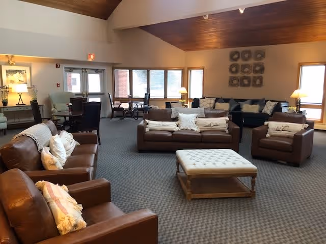 A spacious living room area with multiple brown leather sofas and armchairs arranged around a large cushioned ottoman. The room has a high wooden ceiling, carpeted floor, and large windows letting in natural light. There are side tables with lamps and decorative wall art above one of the sofas.