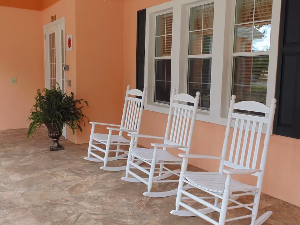 A porch area with three white wooden rocking chairs lined up against a peach-colored wall with three windows. There is a potted green plant near a white door with glass panels.