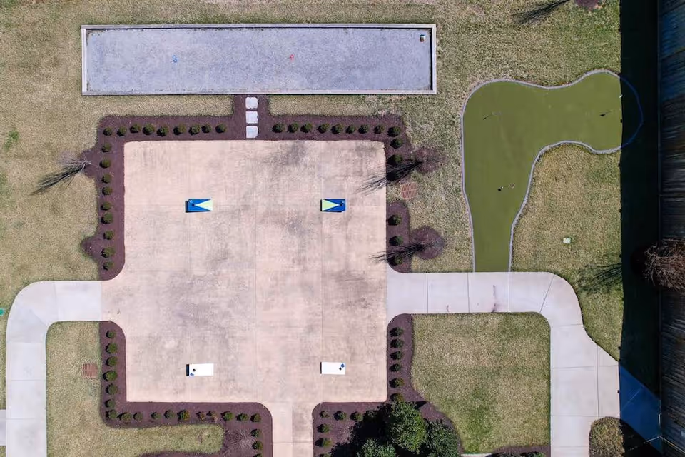 Aerial view of an outdoor recreational area featuring a bocce ball court at the top, a putting green on the right, and a concrete area with two cornhole boards in the center, surrounded by grass and landscaped bushes with paved walkways.