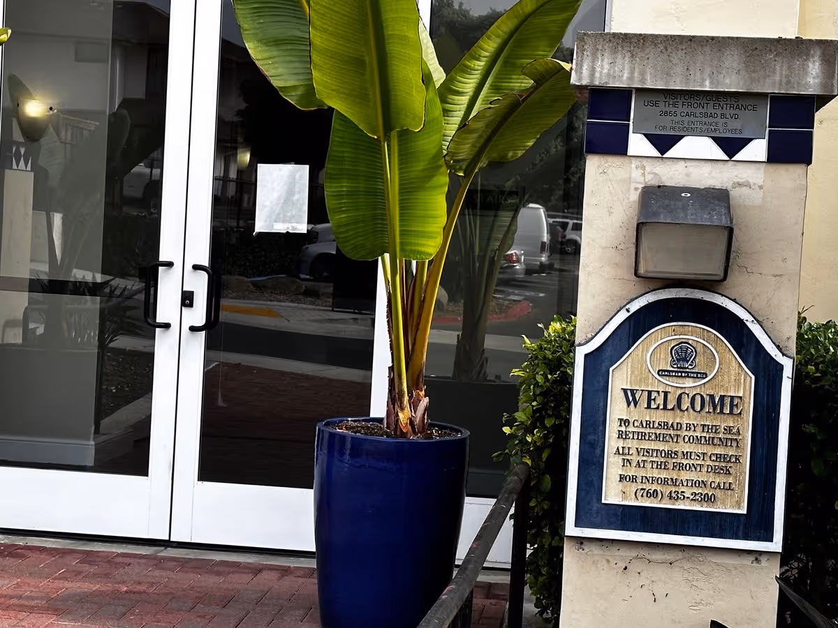 Entrance of Carlsbad By The Sea showing glass double doors, a large potted plant, and a 'Welcome' sign on a pillar.