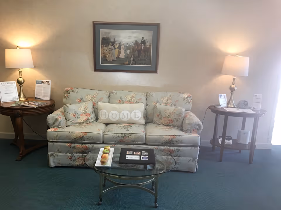 A cozy living room area with a floral-patterned sofa featuring a decorative pillow that spells 'HOME'. On either side of the sofa are wooden side tables with brass lamps and informational pamphlets. A glass coffee table in front of the sofa holds a rectangular white plate with apples and a black photo album. A framed painting depicting a historical scene with people and horses hangs on the wall above the sofa.