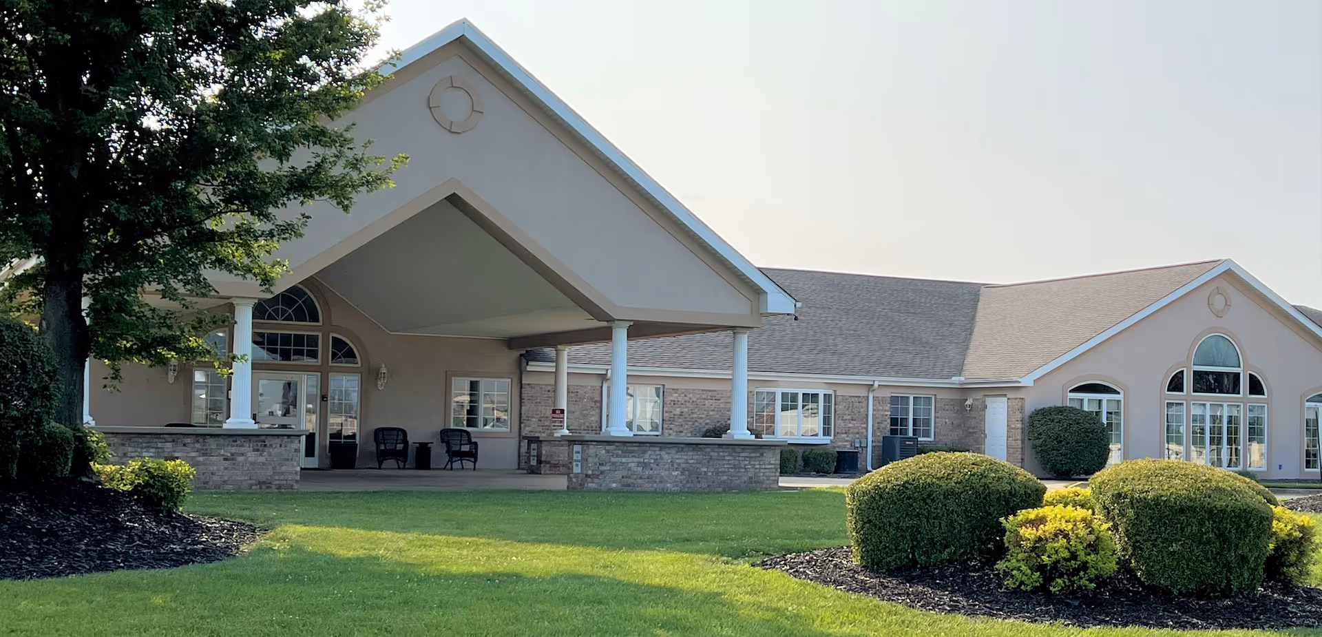 Exterior view of Brookstone Estates of Mattoon South building with a covered entrance supported by white columns, surrounded by green lawn, bushes, and a tree on the left side.