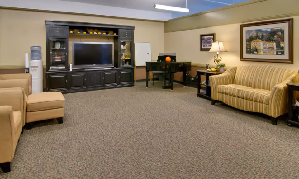 A cozy living room in Heritage Hall featuring a striped sofa, two beige armchairs, a black entertainment center with a large flat-screen TV, a water cooler, a black piano with a small pumpkin on top, a side table with a lamp and decorative items, and framed artwork on the walls.
