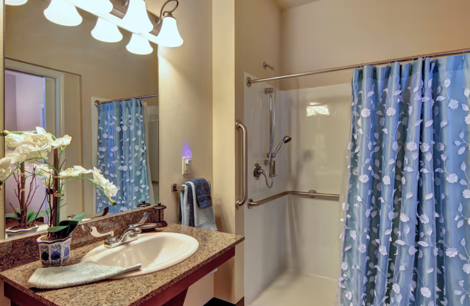 A bathroom with a granite countertop sink, a potted white orchid plant, a towel hanging on a rack, and a shower area with a blue floral shower curtain and grab bars.