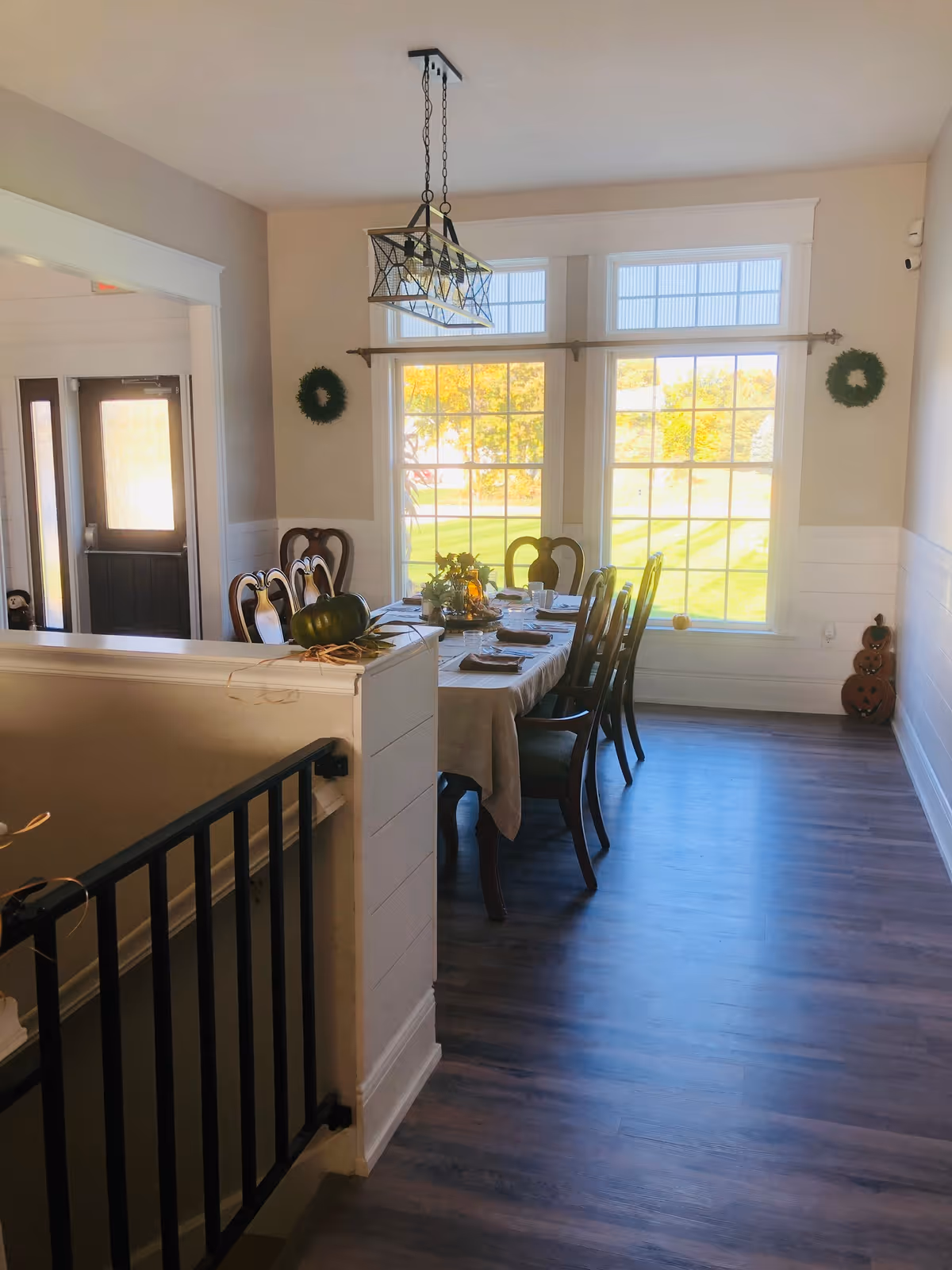 A bright dining room with a long table set for a meal, surrounded by wooden chairs. Large windows let in natural light and show a green outdoor view. The room has light-colored walls, wooden flooring, and decorative wreaths on the walls. A black metal baby gate is visible in the foreground near a staircase.