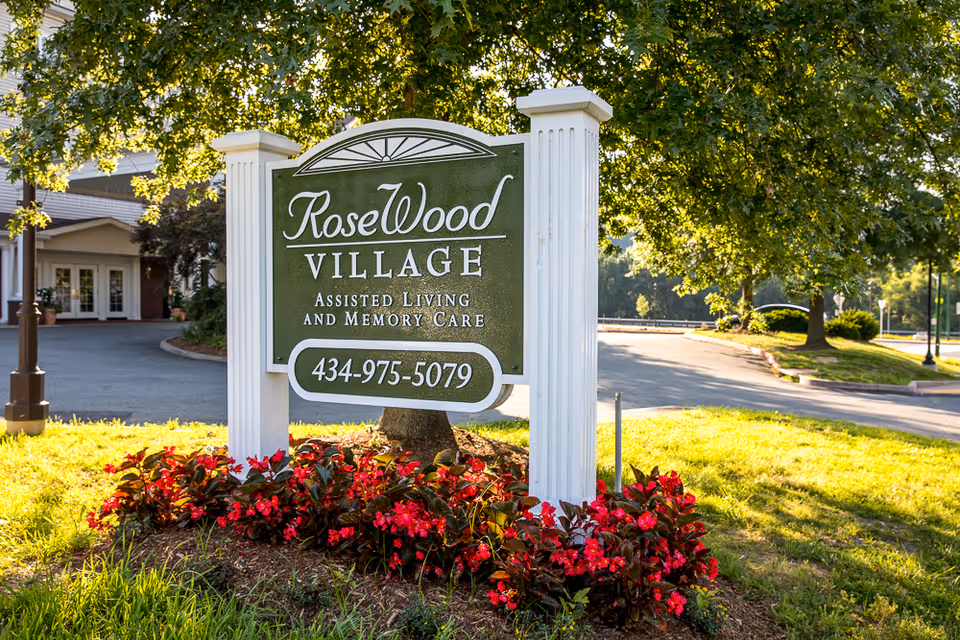 Outdoor view of a green and white sign for RoseWood Village Assisted Living and Memory Care, surrounded by red flowers and greenery, with a building and trees in the background.