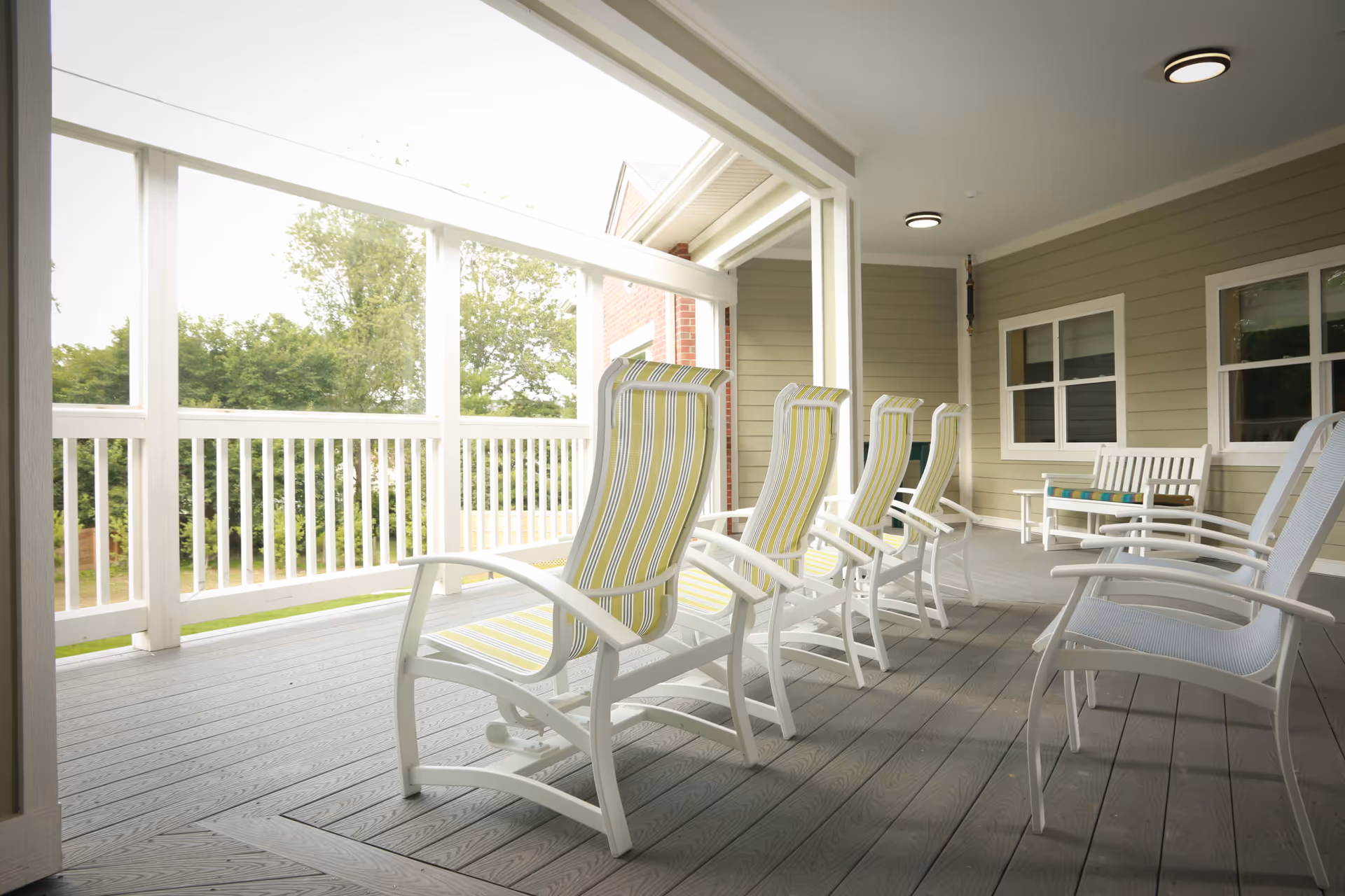 A covered outdoor porch area with several white rocking chairs featuring striped cushions in yellow and green, and a white bench with a colorful cushion. The porch has a gray wooden floor, white railing, and beige siding walls with windows. Trees and greenery are visible outside the porch.