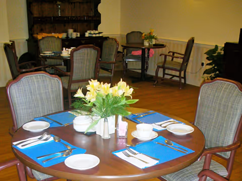 A dining room with round wooden tables set for meals, each table has blue placemats, white plates, cups, and silverware. There is a vase with yellow flowers on the table in the foreground. Several upholstered chairs surround the tables, and the room has wooden flooring and light-colored walls.