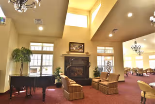 A spacious living room area with high ceilings, featuring a grand piano on the left, two cushioned armchairs facing a fireplace with a framed picture above it, large windows allowing natural light, and a dining area with tables and chairs visible in the background.