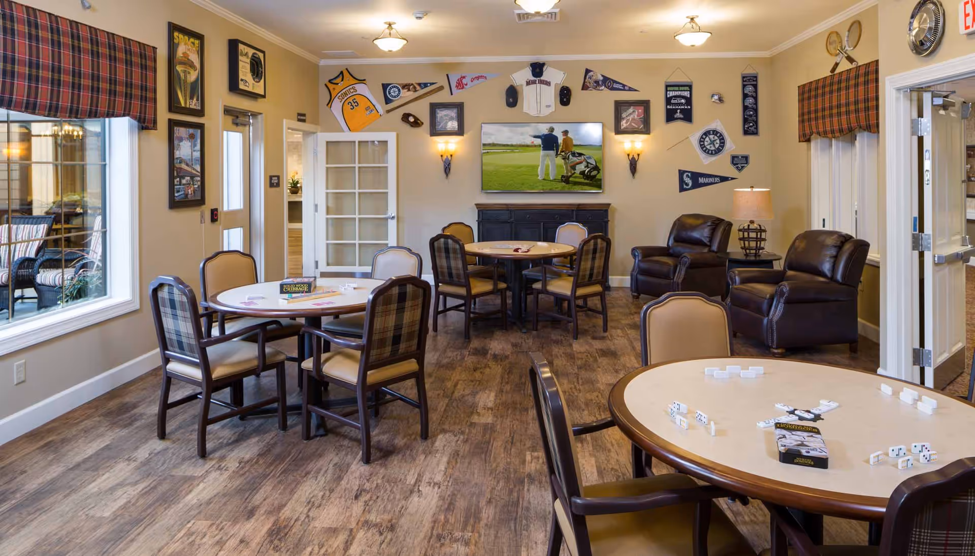 A cozy common room with several round tables and chairs arranged for games, including dominoes and cribbage. The walls are decorated with sports memorabilia such as jerseys, pennants, and framed pictures. There are two leather armchairs near a side table with a lamp, and a flat-screen TV mounted on the wall showing a golf scene. Large windows with plaid valances let in natural light.