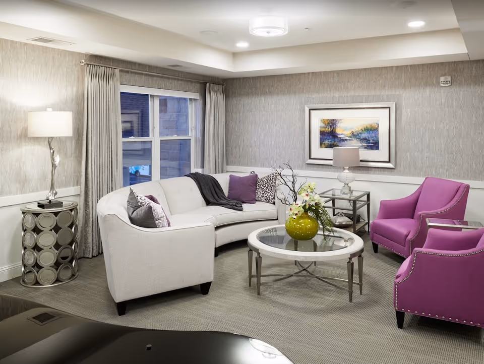 A cozy senior living room with a curved beige sofa adorned with purple and patterned pillows, two bright purple armchairs, a round glass coffee table with a green vase and flowers, a side table with a lamp, and a framed landscape painting on the wall. The room features neutral-toned walls, carpeted floor, and a window with gray curtains.