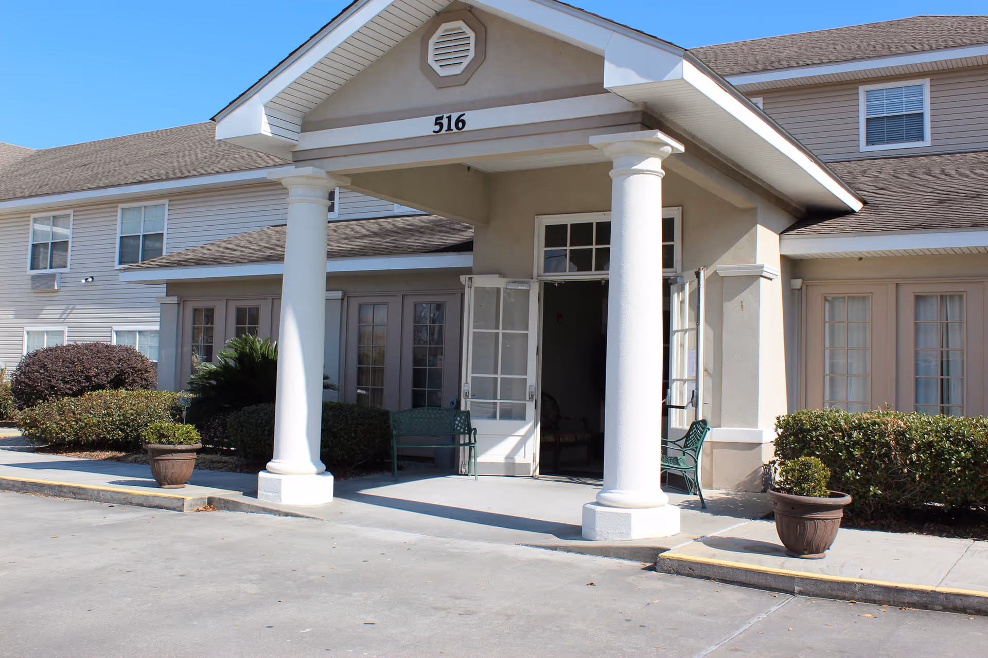 Front entrance of a beige residential building with a covered portico supported by white columns, benches, potted plants, and the number 516 above the doorway.