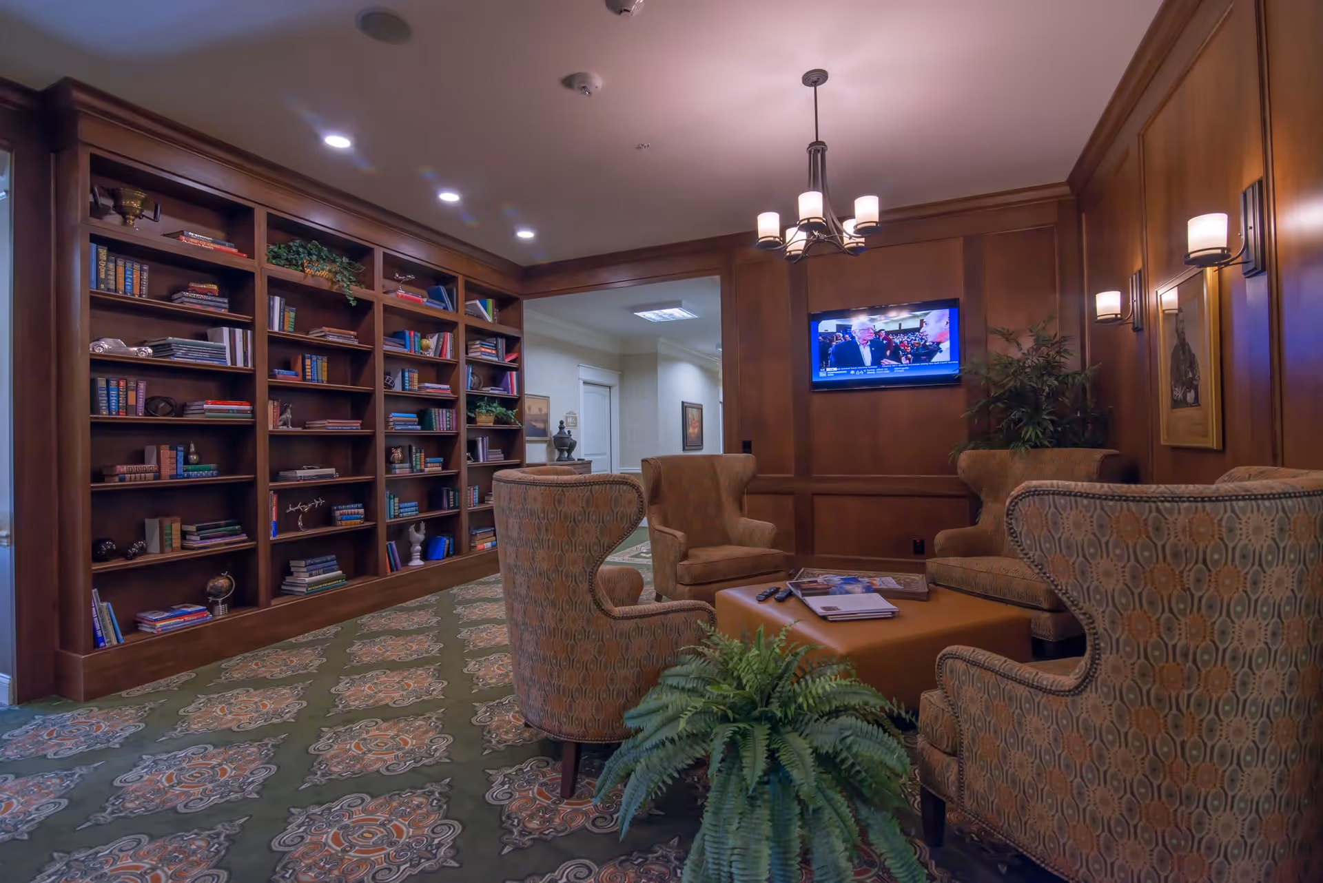 A cozy lounge area with patterned armchairs arranged around a central ottoman, a large built-in wooden bookshelf filled with books and decorative items, a flat-screen TV mounted on a wood-paneled wall, and soft lighting from wall sconces and a ceiling chandelier. A green potted plant is placed in the foreground on a patterned carpet.