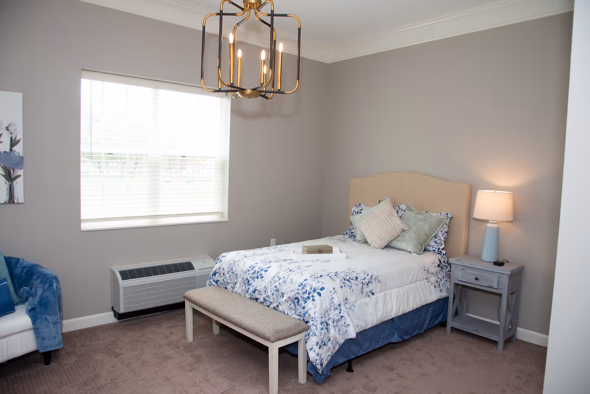 A cozy bedroom with a bed featuring floral bedding and multiple pillows. There is a small bench at the foot of the bed, a nightstand with a lamp on the right side, and a window with blinds letting in natural light. A blue armchair is partially visible on the left side. The walls are painted light gray, and a modern chandelier hangs from the ceiling.