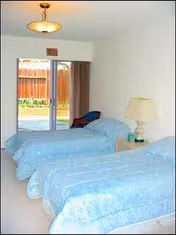 Two twin beds with blue bedding in a bright, minimalist bedroom with a nightstand and lamp.