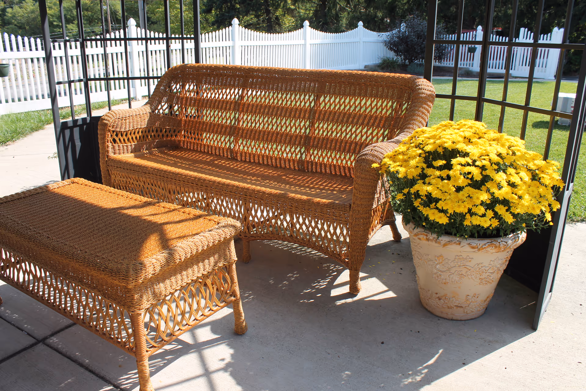 Outdoor seating area with a wicker loveseat and matching wicker table on a concrete patio. Next to the loveseat is a large decorative pot filled with bright yellow flowers. The area is enclosed by a white picket fence and surrounded by green grass and trees.