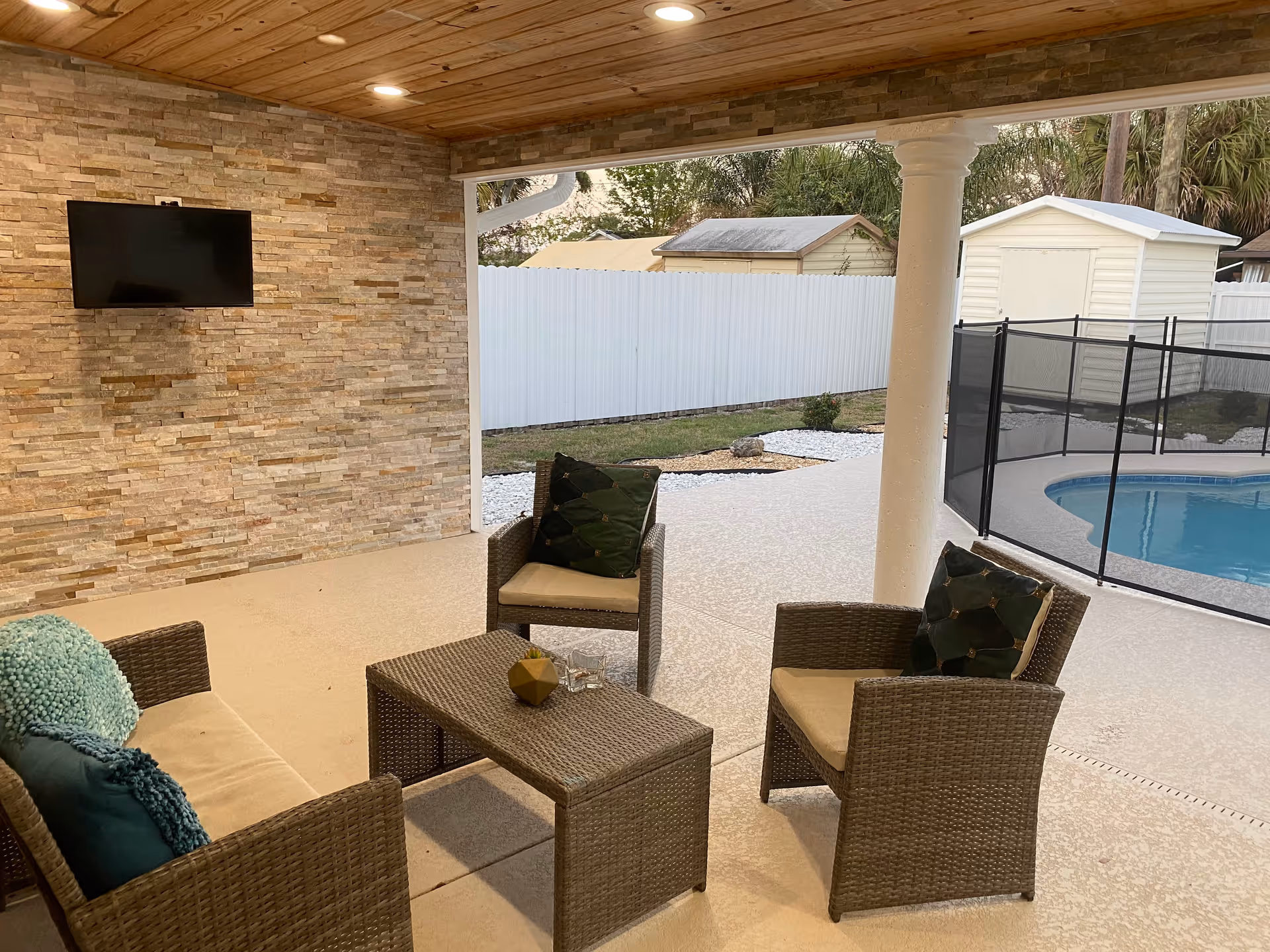 Covered outdoor patio with wicker seating, a coffee table, wall-mounted TV, and a fenced pool visible in the background.