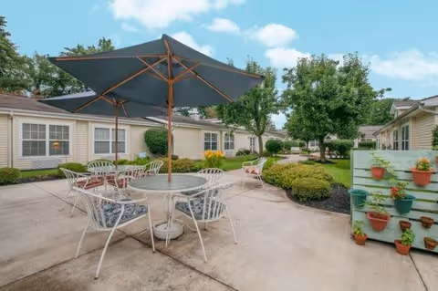Outdoor patio area at The Courtyard at Newark with a round glass table, four white metal chairs with cushions, and a large dark umbrella. The patio is surrounded by well-maintained bushes, trees, and beige single-story buildings under a partly cloudy sky. A vertical garden with potted plants is visible on the right side.
