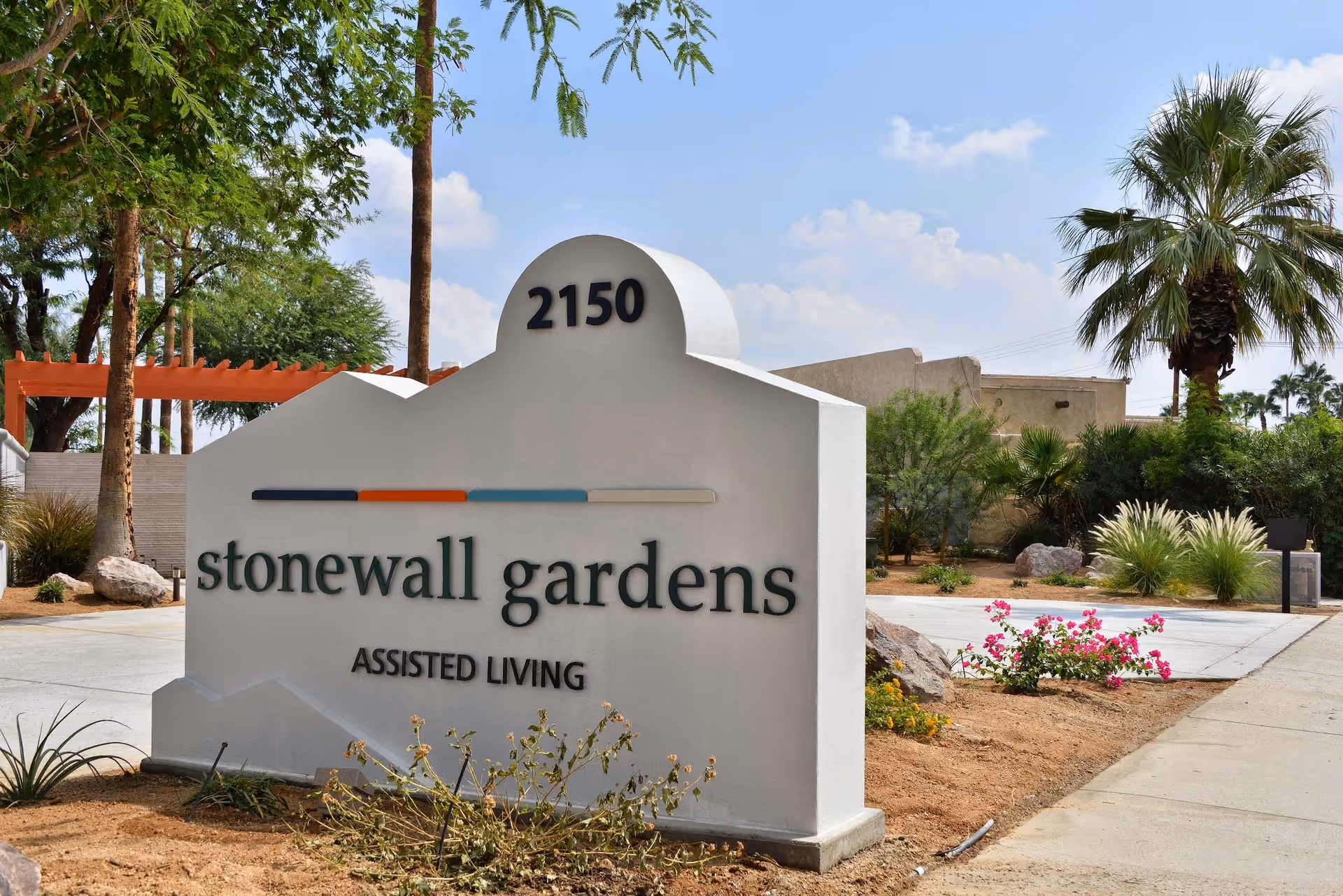 Outdoor view of a white stone sign for Stonewall Gardens Assisted Living with the address number 2150 on top. The sign is surrounded by desert landscaping with small plants, palm trees, and a clear blue sky in the background.