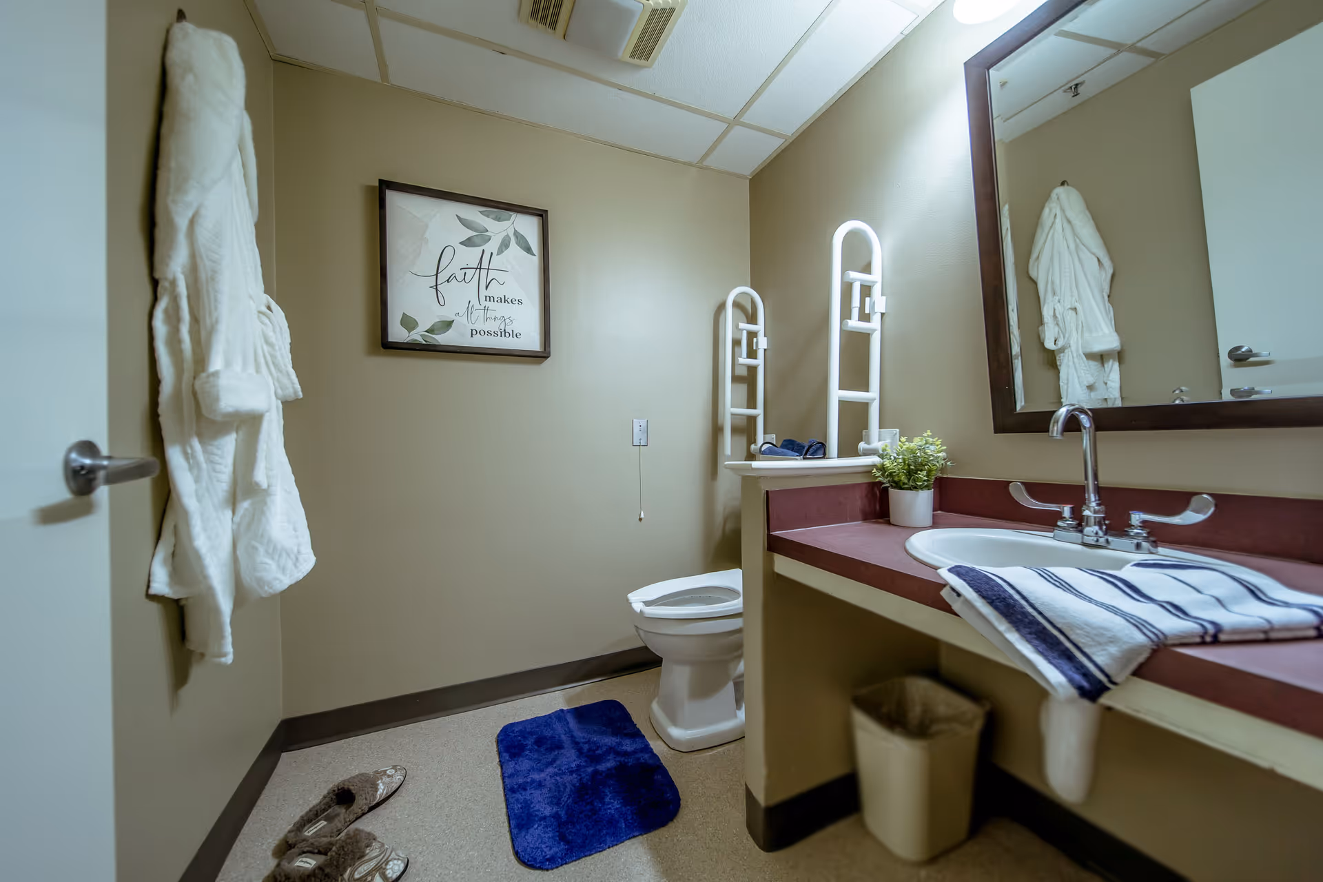 A clean bathroom with beige walls featuring a white toilet, a sink with a maroon countertop, a large mirror, and a white towel hanging on the door. There is a blue bath mat on the floor, slippers nearby, a small potted plant on the counter, and a framed wall art that reads 'faith makes all things possible'.