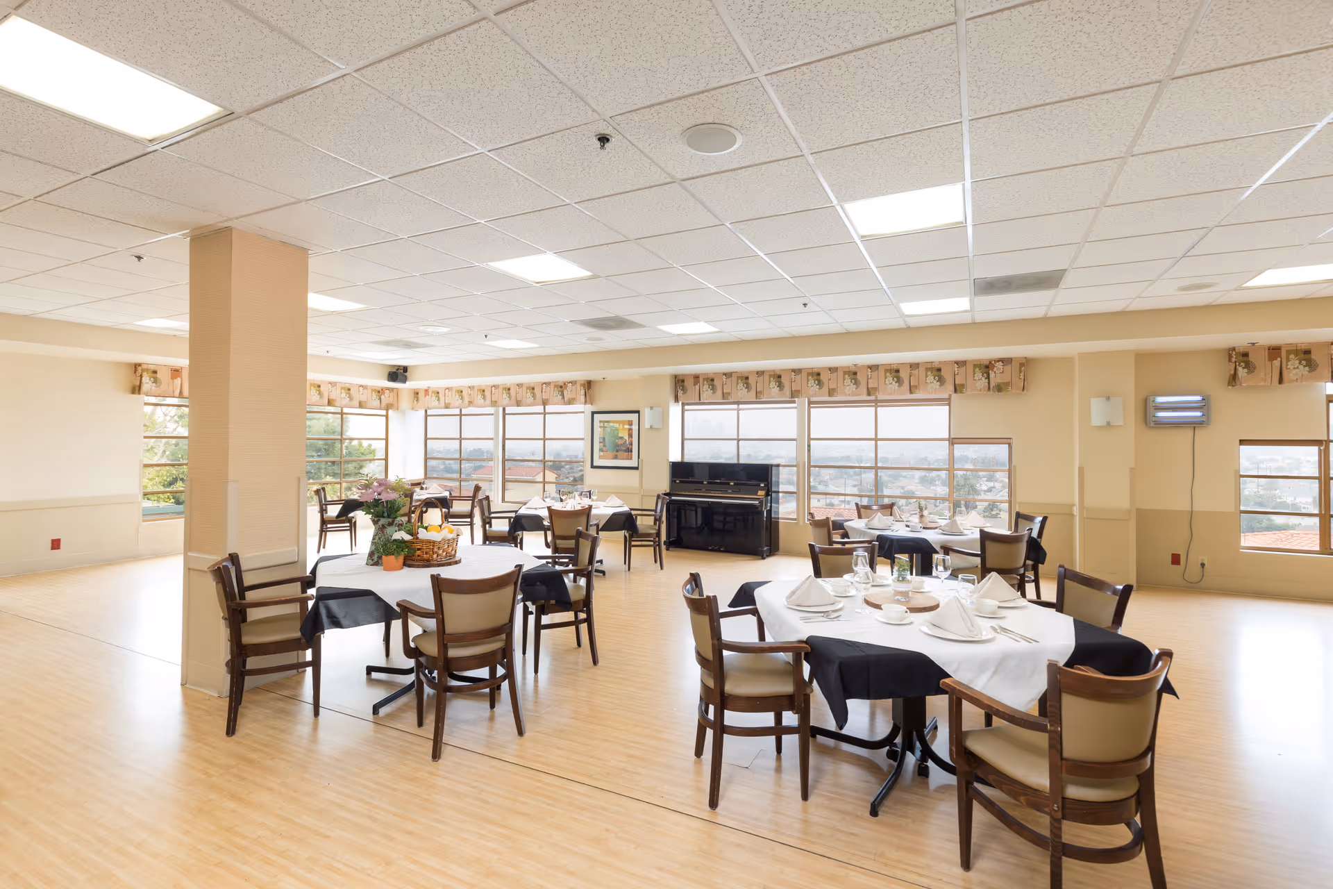 Spacious dining room with round tables set for meals, chairs, large windows and a piano.