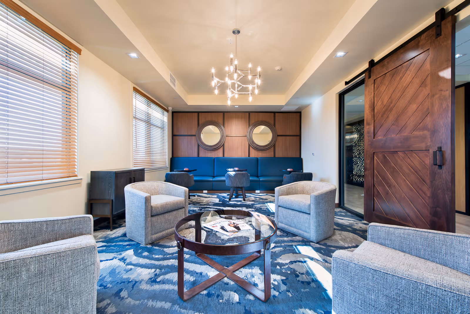 A modern living room with a blue patterned carpet, four gray armchairs arranged around a round glass coffee table with magazines on it, a long blue sofa against a wooden paneled wall with two round mirrors, large windows with wooden blinds, a contemporary chandelier hanging from the ceiling, and a large wooden sliding barn door on the right.