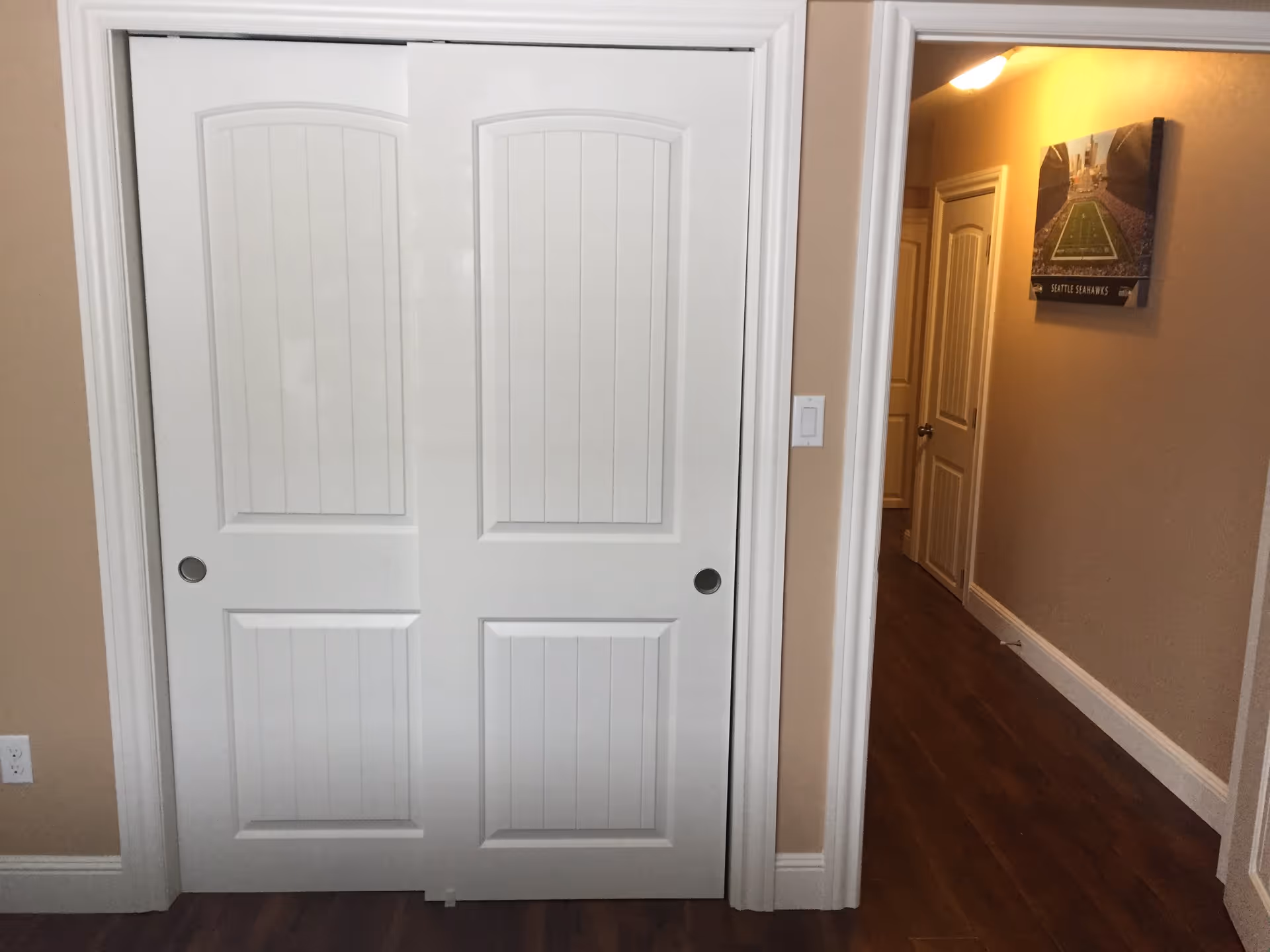 Interior view of a hallway with wooden flooring and beige walls. There are two white sliding closet doors on the left side and a hallway leading to other rooms on the right. A framed picture of a Seattle Seahawks football field is hung on the wall in the hallway.
