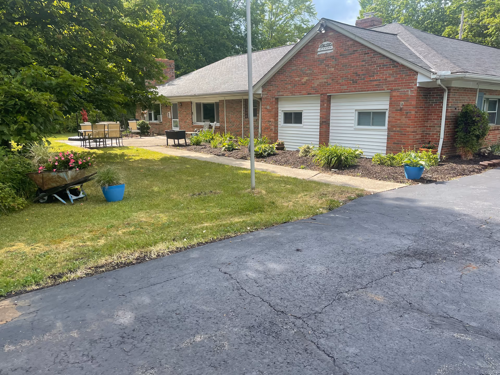 Exterior view of a single-story brick building with a gray roof, two white garage doors, and a paved driveway. The building is surrounded by green grass, plants, and trees. There is outdoor patio furniture on a concrete patio area to the left of the building.
