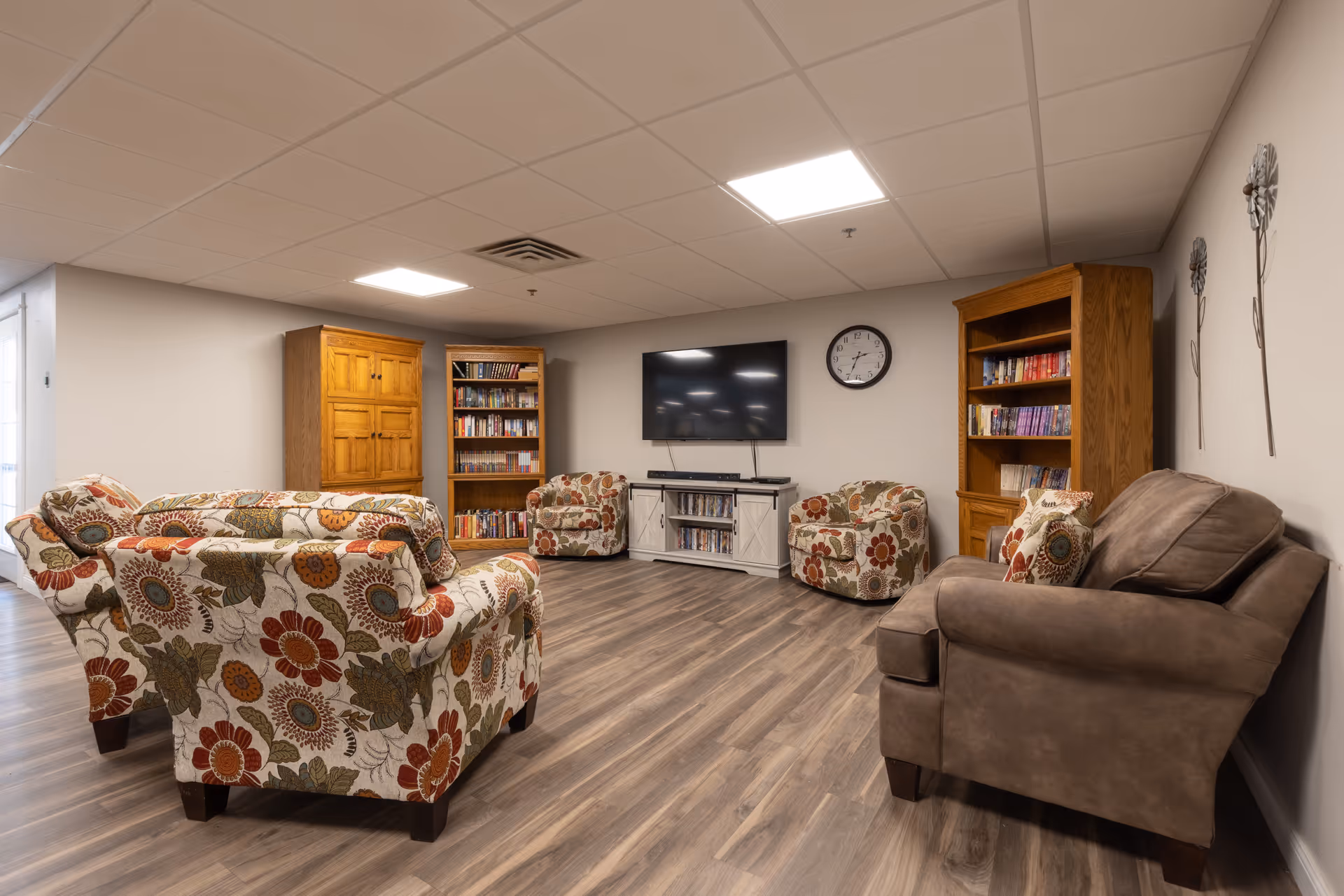 Cozy common living area with patterned armchairs, a TV on a media console, bookshelves, and wood-look flooring.