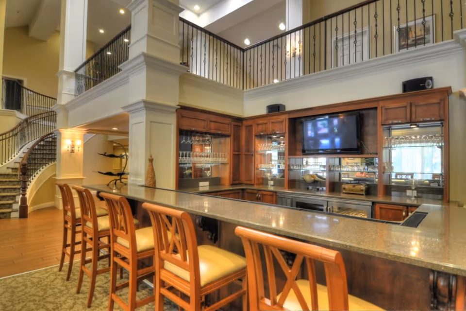 Interior view of a senior living facility bar area with a long countertop and five wooden bar stools with cushioned seats. Behind the bar is a wooden cabinet with glass shelves holding glassware, and a flat-screen TV mounted on the wall. To the left, a carpeted staircase with a wooden handrail curves upward to a second-floor balcony with black wrought iron railings. The space is well-lit with recessed ceiling lights and wall sconces.