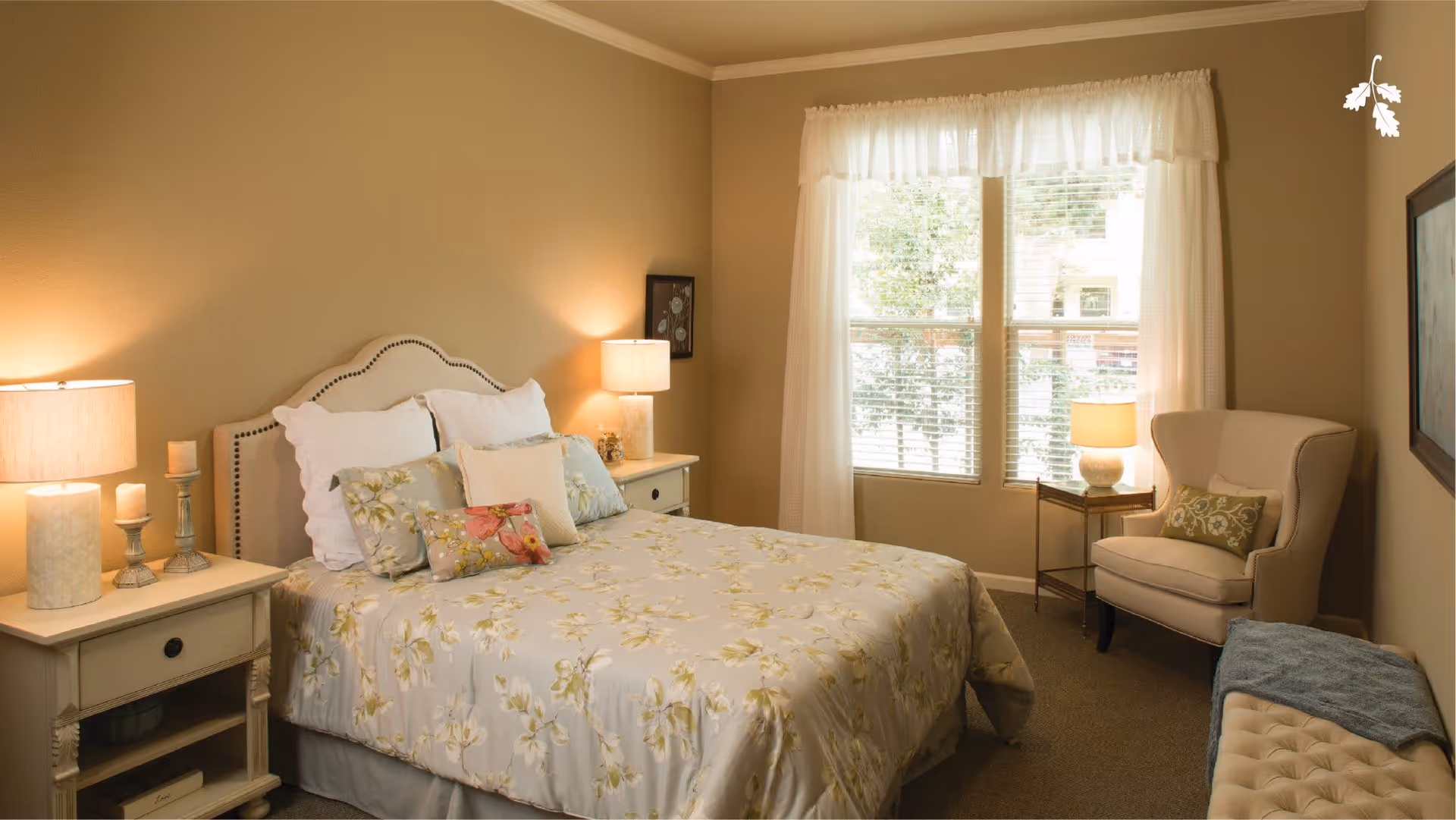A cozy bedroom with a bed covered in floral bedding and multiple pillows. There are two nightstands on either side of the bed, each with a lamp and decorative items. A window with sheer white curtains lets in natural light. A cushioned armchair with a decorative pillow sits near the window, and a bench with a folded blanket is placed against the opposite wall. The walls are painted beige, and the room has a warm, inviting atmosphere.