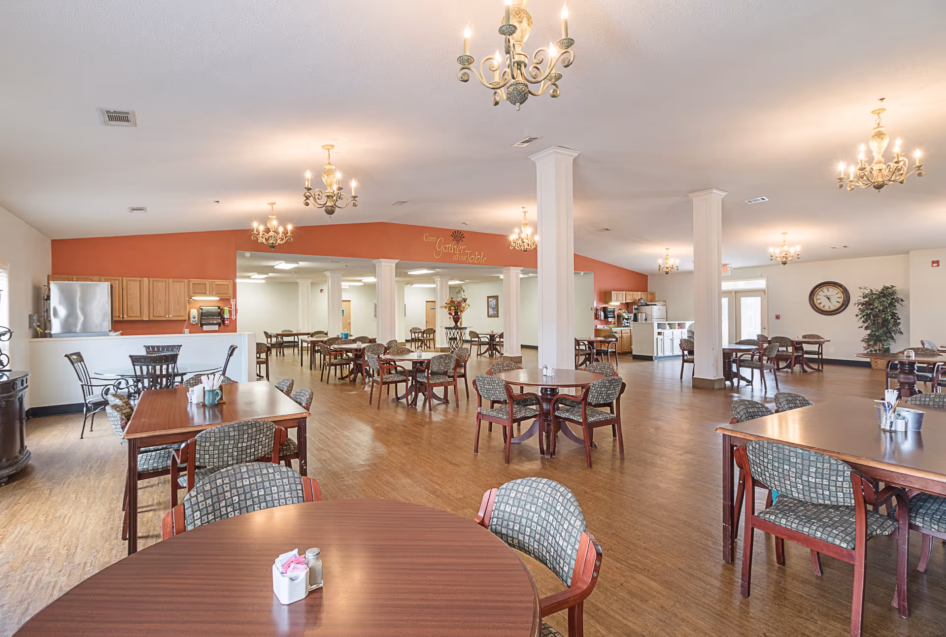A spacious dining room with multiple wooden tables and cushioned chairs arranged neatly. The room features hardwood flooring, several chandeliers hanging from the ceiling, and a kitchen area with cabinets and a refrigerator in the background. A decorative sign on the wall reads 'Come Gather at our Table'.
