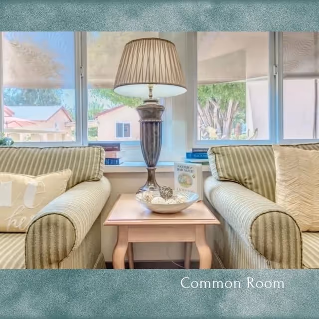 A cozy common room with two striped armchairs facing each other, separated by a wooden side table holding a decorative bowl and a lamp. Behind the table is a window with a view of houses and trees outside.