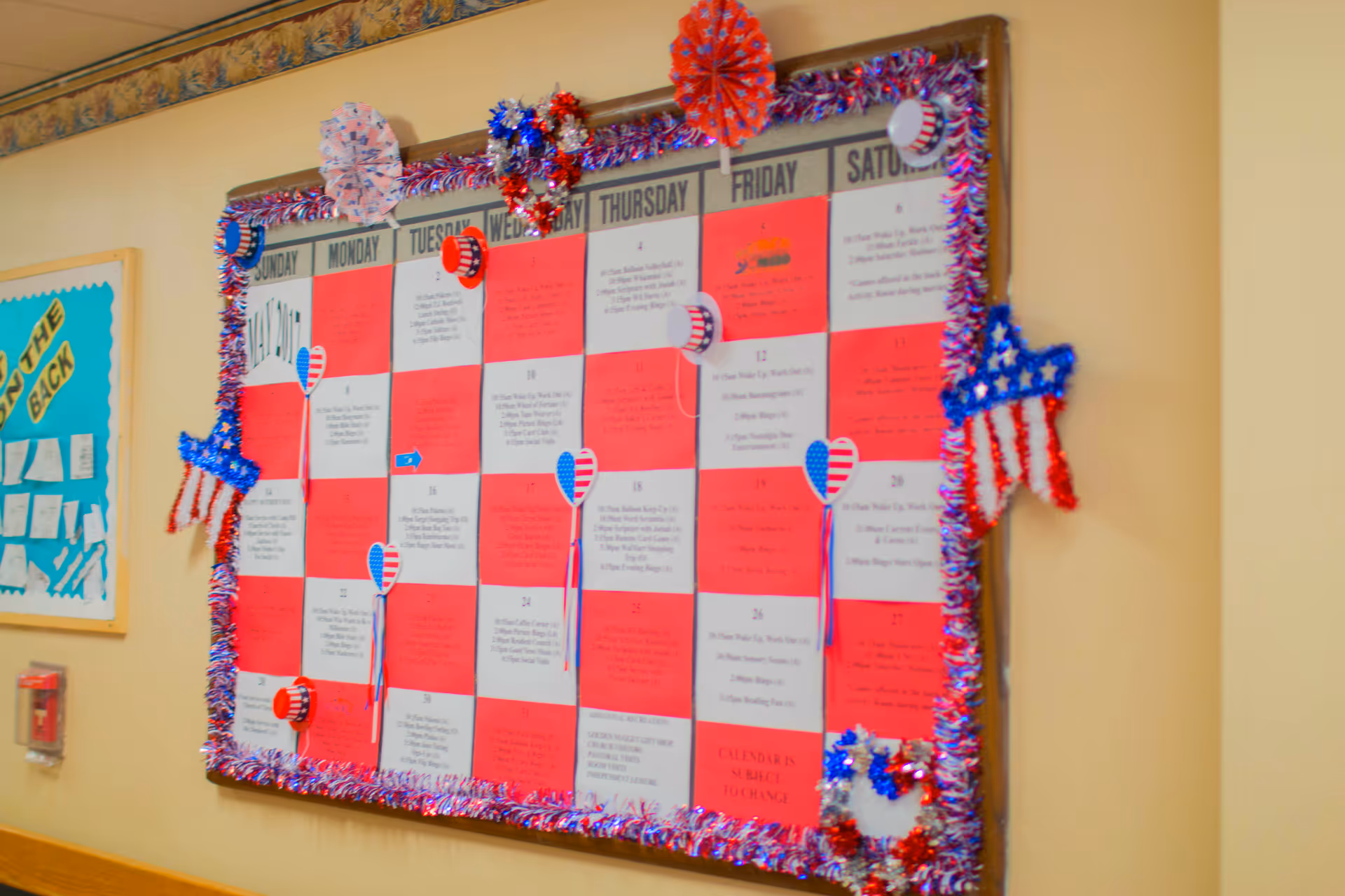A large bulletin board calendar decorated with red, white, and blue patriotic decorations including stars, hearts, and hats. The calendar displays the days of the week and various scheduled activities. The board is framed with red, white, and blue tinsel garland and is mounted on a beige wall.