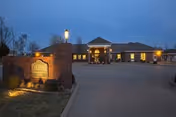 Evening view of Oak Manor Nursing Center showing a well-lit brick entrance sign on the left and the main building with illuminated windows and entrance in the background under a darkening sky.