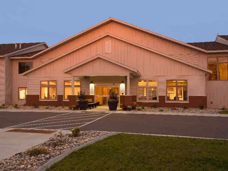 Front entrance of a single-story senior living building with a covered porch, lit windows, planters, and a small parking area at dusk.
