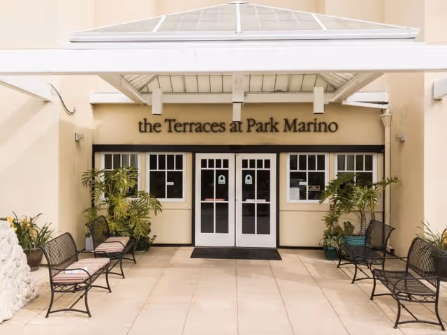 Entrance to The Terraces at Park Marino facility with double glass doors, a covered awning, potted plants, and benches with cushions on either side of the walkway.